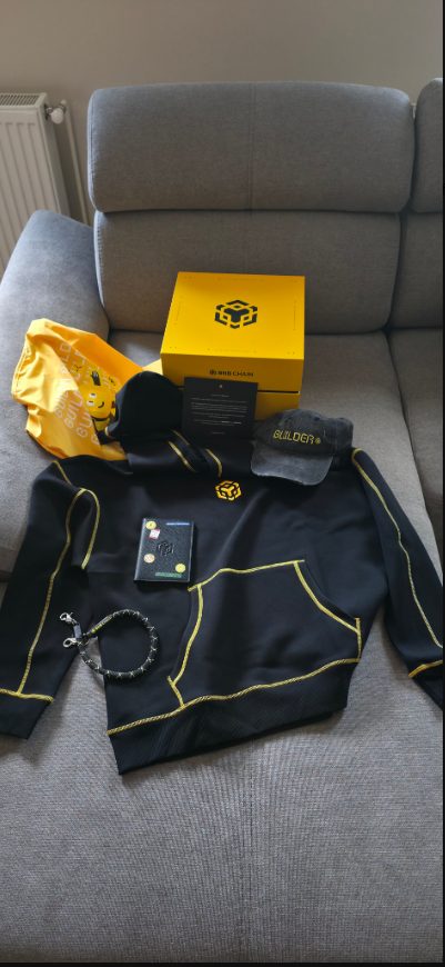 Full BNB Chain anniversary box contents laid out: yellow box, hoodie, cap, card, and accessories