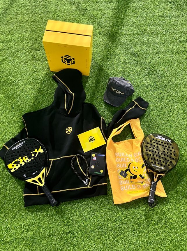 The BNB Chain anniversary box unboxed: hoodie, cap, tote bag, card holder, stickers, and padel rackets on grass